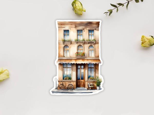 Sticker eines zweistöckigen Geschäfts im mediterranen Stil mit Balkon und Blumenkästen an den Fenstern. Vor dem Geschäft steht ein Fahrrad und ein kleiner Tisch mit Pflanzen. Die Fassade ist in warmen Gelbtönen gehalten.